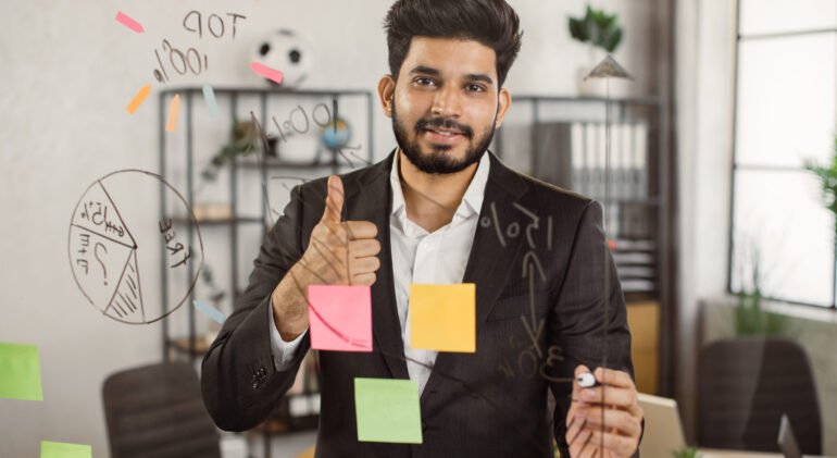 Portrait of executive male manager showing thumb up sign while working on new project at office. Smiling indian man standing in front of glass board and sharing with motivation mood.